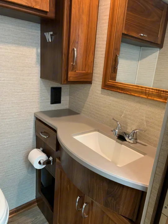 This RV rental's compact, well-equipped bathroom features a vanity with sink, faucet, mirror, wooden cabinets, and toilet paper.. Winnebago Outlook 2020