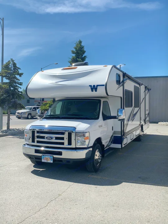 White Ford Class C RV, possibly an rv rental, parked on asphalt. Features an Alaska license plate (JUD 799) on a sunny day.. Winnebago Outlook 2020