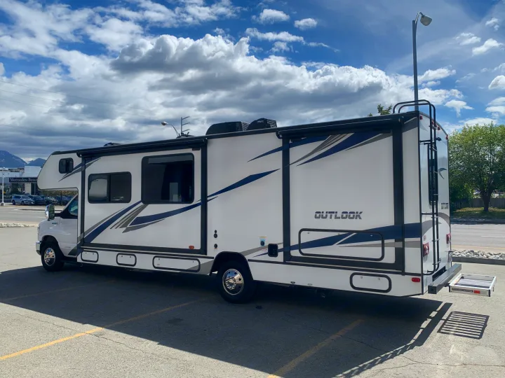 A white "OUTLOOK" Class C motorhome rv rental is parked outdoors under a blue, cloudy sky, showcasing its side and rear.. Winnebago Outlook 2020