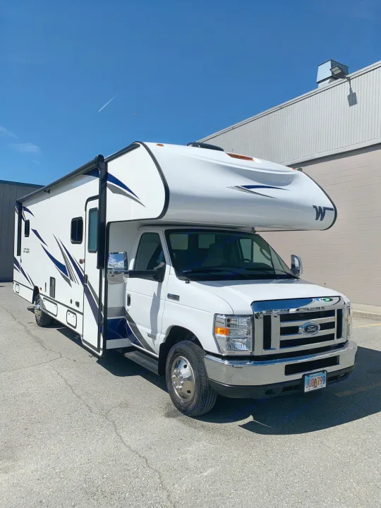 White Winnebago Ford Class C rv rental with blue graphics, parked on asphalt under a clear sky. Ready for your travel adventure.. Winnebago Outlook 2020