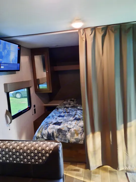 Inside an RV rental, a bunk area with a privacy curtain, patterned bedding, a small mirror, a TV, and a window view.. Dutchmen Coleman Lantern LT 2020