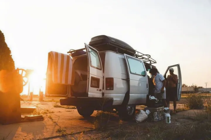 Enjoy an RV rental adventure! Two men organize their camper van at sunset, gear piled outside. Ideal for travel.. Ford Ford E350 2011