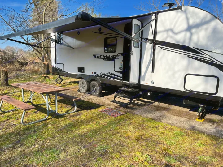 Sunny day at an RV rental campsite. This Prime Time Tracer travel trailer has its awning extended over a picnic table, ready for camping.. Forest River Tracer 2022