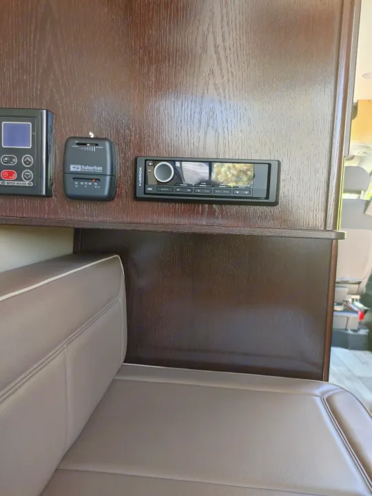 RV rental interior showing water heater, Suburban thermostat, and Furrion entertainment system mounted on wood grain paneling above seating.. Regency RV National Traveler 2019