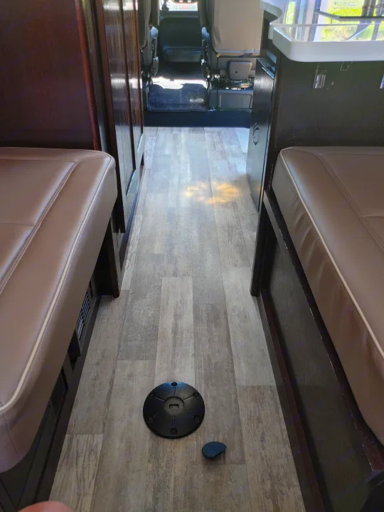 RV rental interior: A narrow aisle with light wood-look floor, flanked by taupe seats & dark cabinetry. Table base on floor, leads to vehicle cab.. Regency RV National Traveler 2019