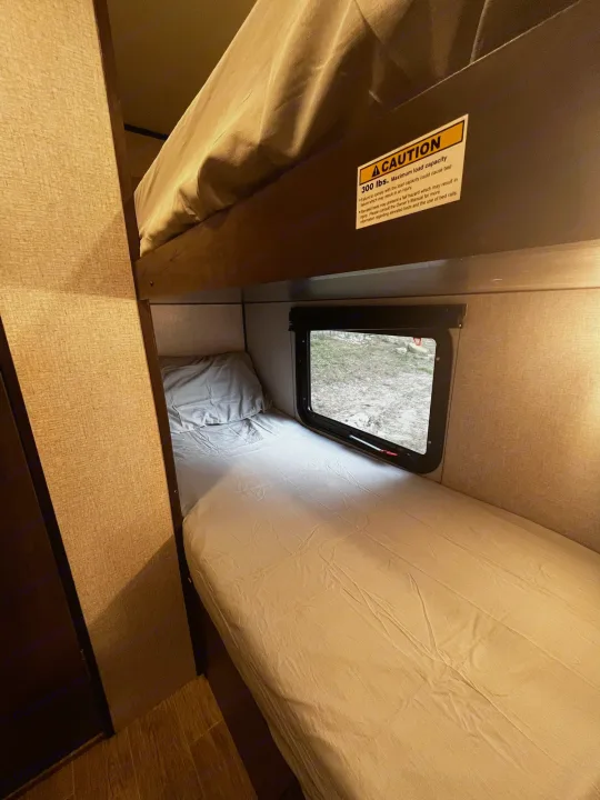 Inside an RV rental: cozy bunk beds with a safety warning (300 lbs capacity) and a window view. Ideal for campers.. Coleman CM17B21 2021