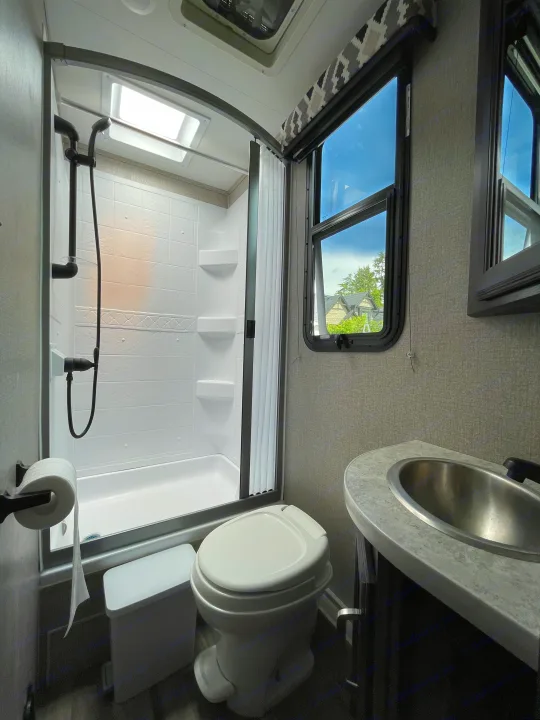 RV rental bathroom featuring a white shower, toilet, and stainless sink. Bright windows show sky & trees. Functional space.. Jayco Redhawk 2021