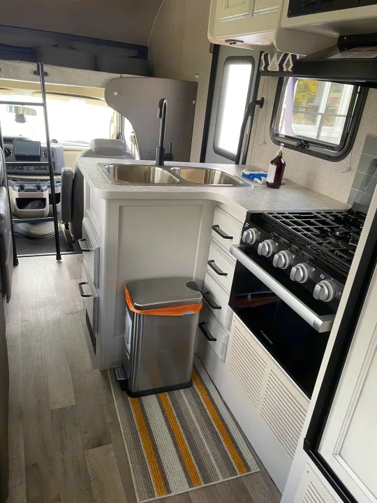 RV rental interior: Modern kitchen with Furrion stove, double sink, wood-look floor, and striped rug.. Jayco Redhawk 2021