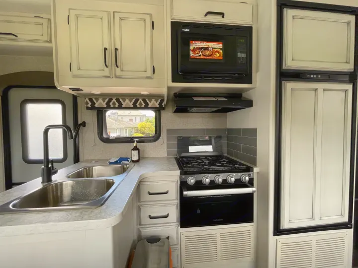 Well-equipped modern RV rental kitchen featuring a double sink, black faucet, microwave, gas stove, and refrigerator.. Jayco Redhawk 2021
