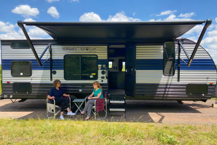 Two women relax in camping chairs next to a large Grey Wolf RV rental with its awning extended on a sunny day.. Forest River Cherokee Greywolf 2022