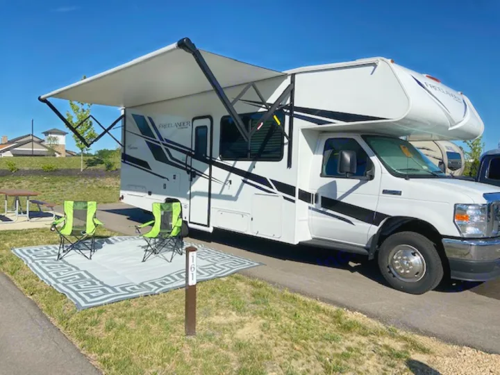 White Class C RV rental set up at a sunny campsite with extended awning, two green chairs, and an outdoor rug.. Forest River Coachmen Freelander Premier 2022