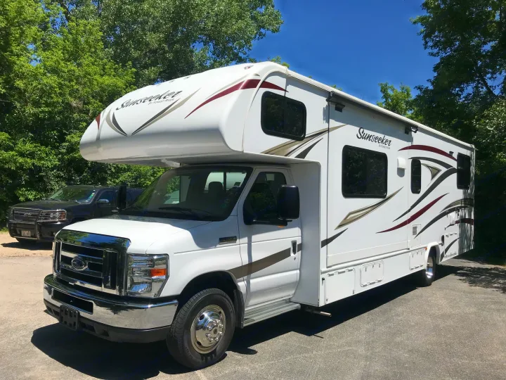 Bright white Sunseeker Class C RV rental, ready for travel. Parked on asphalt with green trees and blue sky.. Forest River Sunseeker 2017