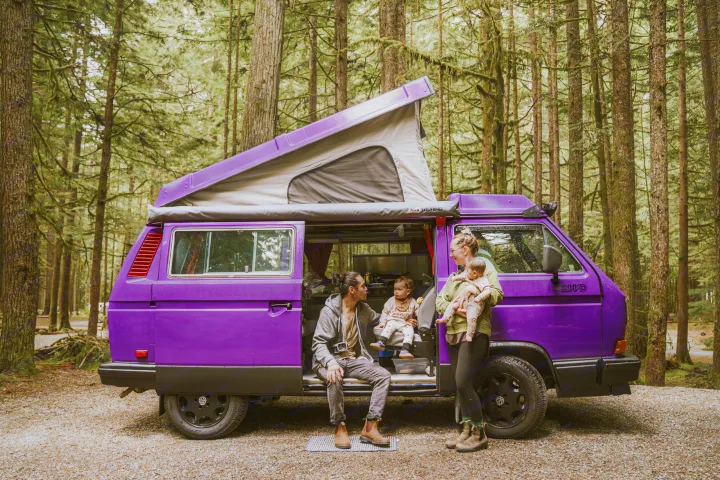 A family with two children enjoys their purple pop-top camper van, an RV rental, at a lush forest campsite.. Volkswagen Vanagon 1991