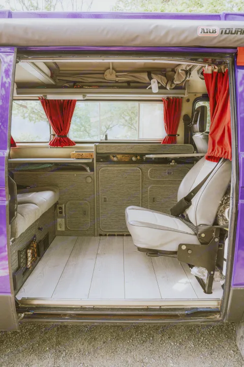 An RV rental's inviting interior: light wood floor, cozy grey seating, kitchen area, and red curtains.. Volkswagen Vanagon 1991