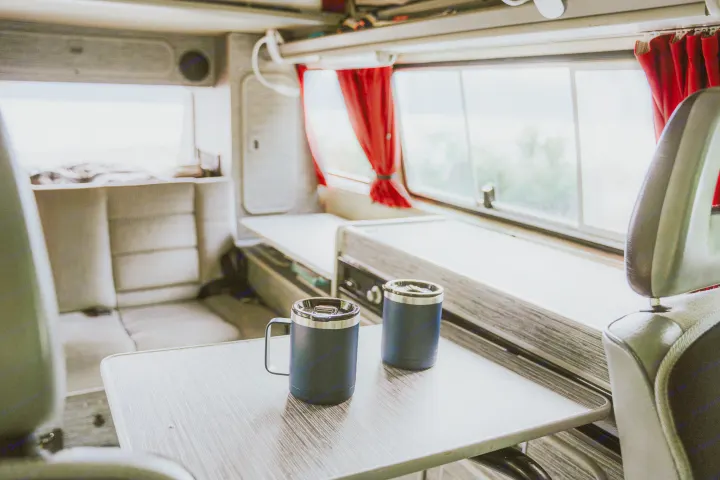 Cozy RV rental interior: table with two blue travel mugs, comfy seating, & bright window views. Perfect for adventure.. Volkswagen Vanagon 1991