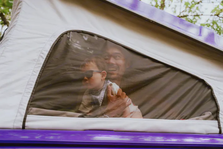 Smiling dad and child relaxing inside their RV rental's rooftop tent. Purple vehicle base visible.. Volkswagen Vanagon 1991