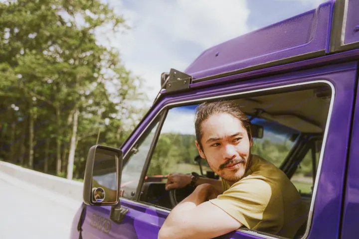 A man with a mustache leans out a vibrant purple camper van, ready for your next RV rental adventure.. Volkswagen Vanagon 1991