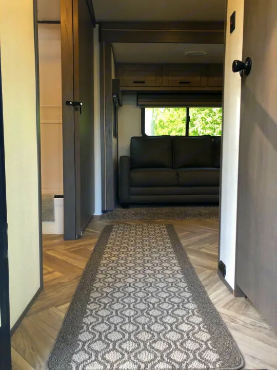 Inside your RV rental: Hallway perspective showing living room with a black sofa, overhead cabinets, and bright window views.. Cruiser Rv Corp Shadow Cruiser 2021