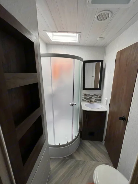 Inside an RV rental bathroom: corner shower, vanity with sink, mirror, toilet, and built-in storage shelves. Compact and functional space.. Forest River Shockwave 2022