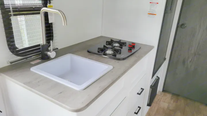 Compact RV rental kitchen features a white sink, modern faucet, two-burner cooktop, and gray counter with white cabinets.. Keystone Hideout 2022
