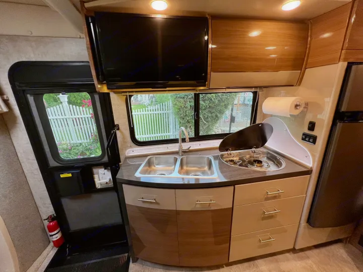 Modern RV rental kitchen featuring a sink, stovetop, TV, cabinets, and a side door with a view of a picket fence.. Itasca Navion 2015