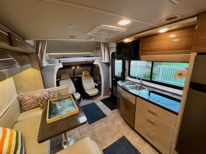 RV rental interior: Modern layout with a bright dinette, functional kitchen, and bunk bed. Windows show outdoor view.. Itasca Navion 2015