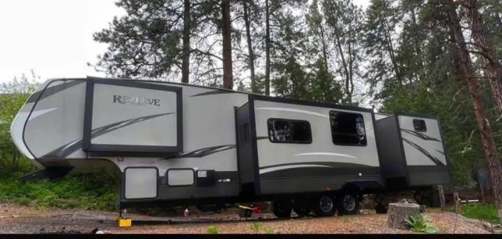 A large, light gray fifth-wheel RV rental with extended slide-outs is parked in a treed outdoor setting.. Crossroads RV Rezerve 2016