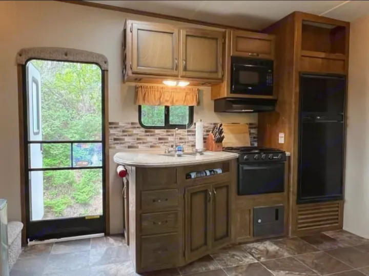 RV rental kitchen with sink, stove, microwave, fridge, and cabinets. The entry door provides a view of the surrounding trees.. Crossroads RV Rezerve 2016