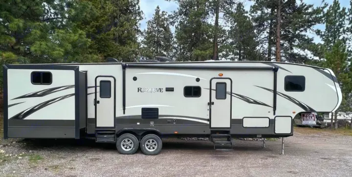 A long, white and dark grey fifth-wheel RV, common for rv rental, is parked with its slide-outs extended. Pine trees frame this travel home.. Crossroads RV Rezerve 2016