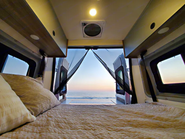 Cozy rv rental bed view: Ocean sunset through open back doors with mesh screens from inside a camper van.. Winnebago Solis 2023