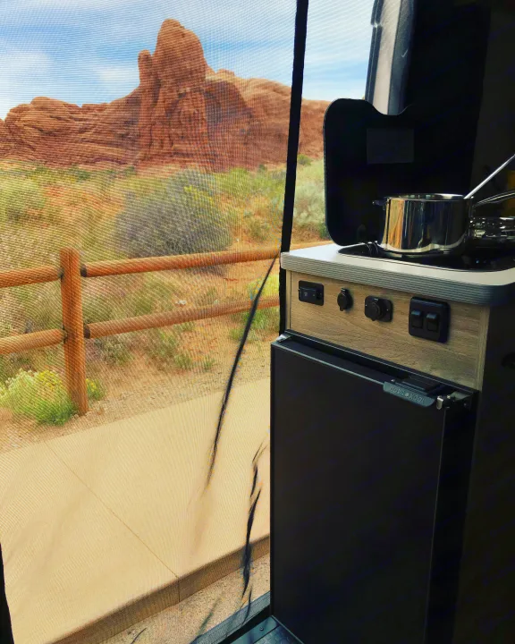 RV rental interior showing a compact camper kitchen, with a stunning desert view of red rock formations through a mesh screen.. Winnebago Solis 2022