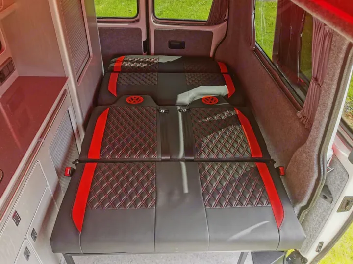 Inside an RV rental: Convertible bed/seat setup in a campervan. Features red & black quilted upholstery & VW logo.. Volkswagen T6 Transporter LWB 2018