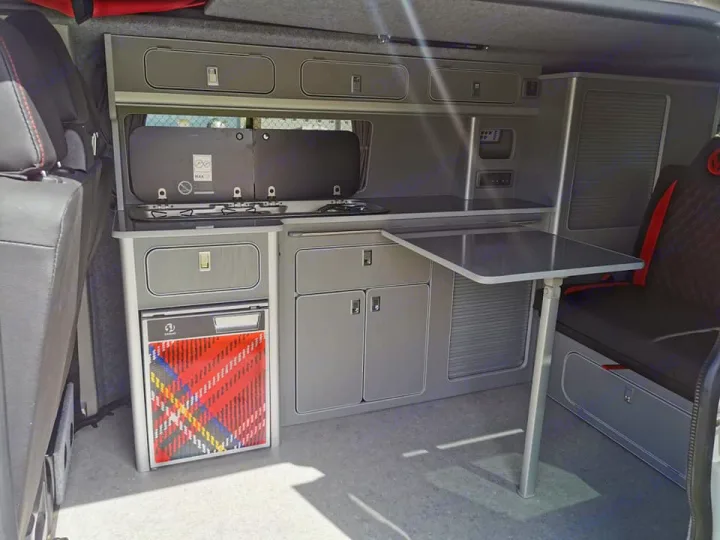 Well-equipped RV rental showing a grey camper van interior with a kitchen, plaid fridge, storage, and fold-down table.. Volkswagen T6 Transporter LWB 2018