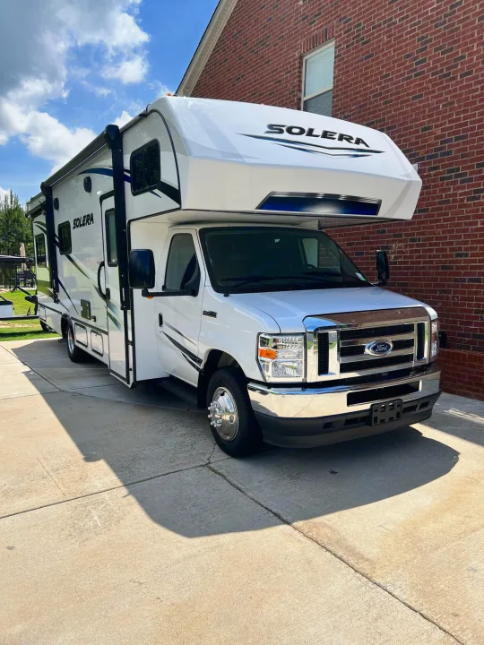 A white Solera Class C RV rental is parked on a driveway beside a brick house under a blue sky, ready for adventure.. Forest River Solera 2022