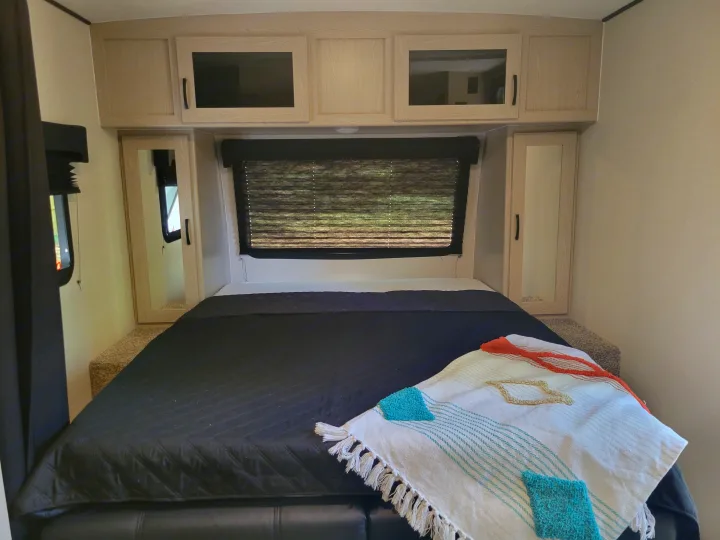 This RV rental bedroom offers a comfortable bed with dark comforter & colorful throw, surrounded by practical light wood cabinets.. Other Other 2022