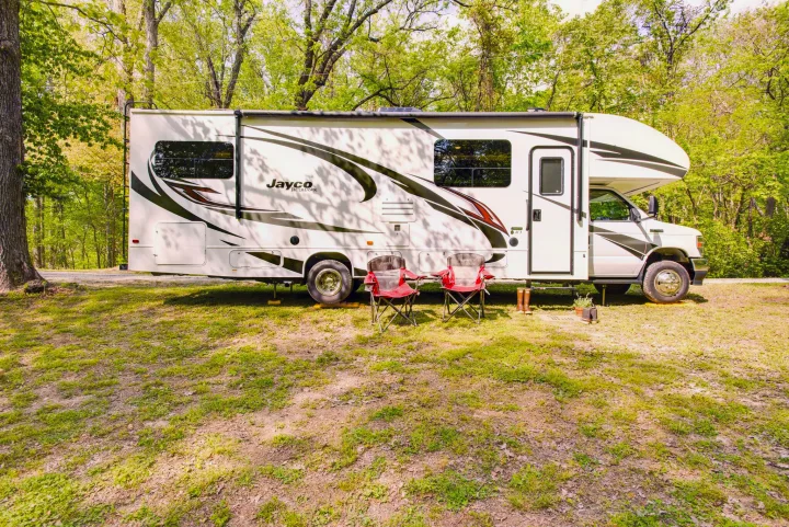 Jayco Redhawk RV rental parked at a sunny campground. Two red chairs are set up in front of the motorhome amidst trees.. Jayco Redhawk 2023