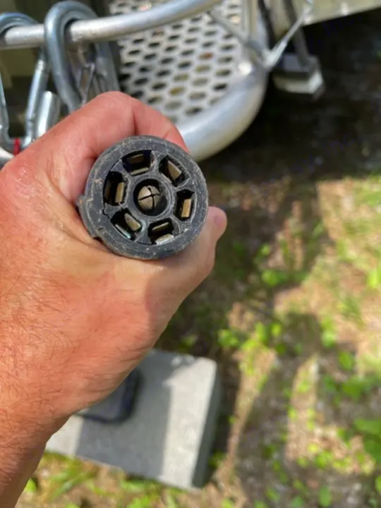 A hand holds a 7-pin RV trailer electrical plug, essential for rv rental and towing. Visually impaired: Round black connector.. NuCamp T@G MAX XL BOONDOCK 2021