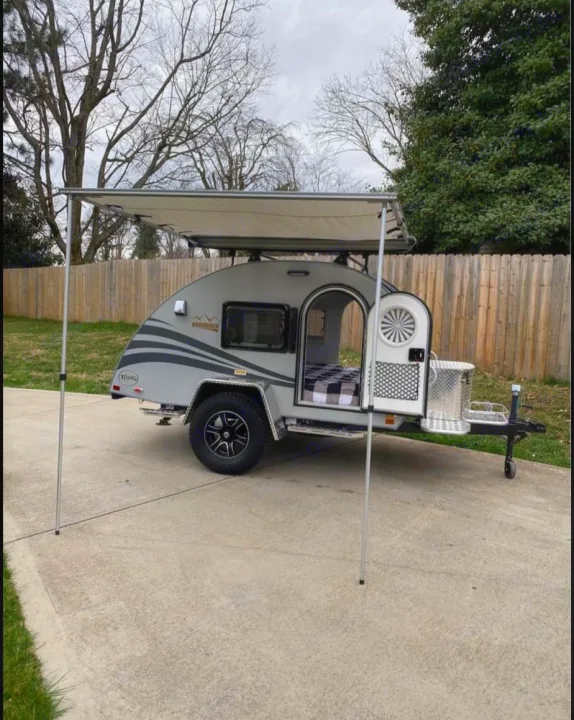 Exterior view of a grey teardrop RV rental with an extended awning. The open door reveals a cozy checkered bed inside.. NuCamp T@G MAX XL BOONDOCK 2021