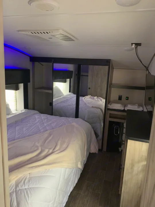 Inside an RV rental, a bedroom with white bedding, blue LED lights, ceiling vents, and mirrored closets reflecting the bed.. Forest River Cherokee Arctic Wolf 2022