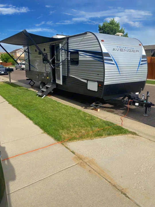 An RV rental, a gray Prime Time Avenger travel trailer, is parked curbside with its awning extended and steps down, ready for use.. Prime Time Avenger 2021