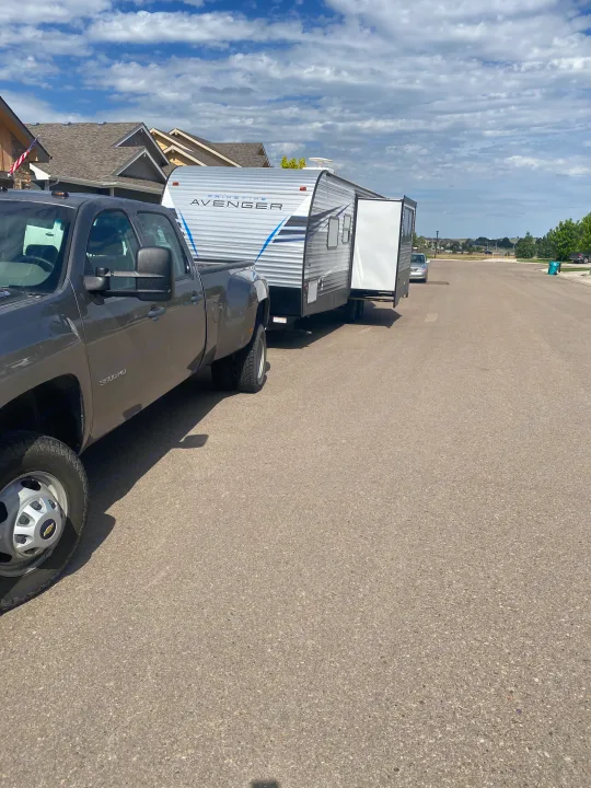 Grey Chevy 3500 HD pickup towing a Prime Time Avenger RV rental on a sunny residential street with houses.. Prime Time Avenger 2021
