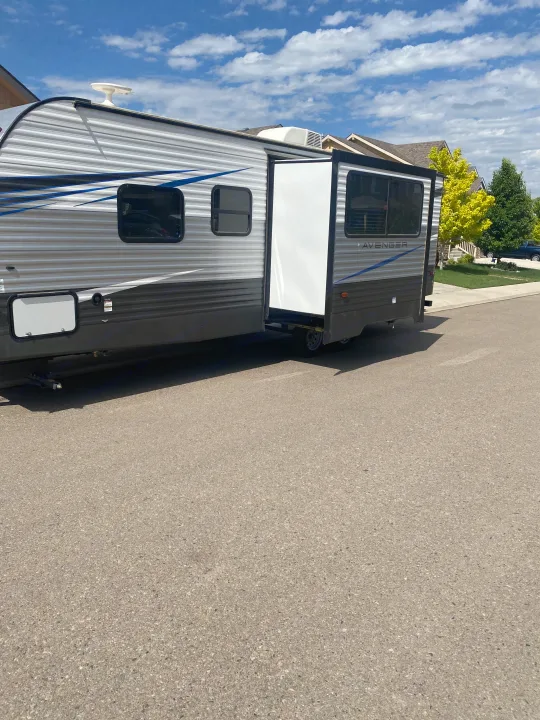 This rv rental, a travel trailer with an extended slide-out, is parked on a residential street on a sunny day.. Prime Time Avenger 2021