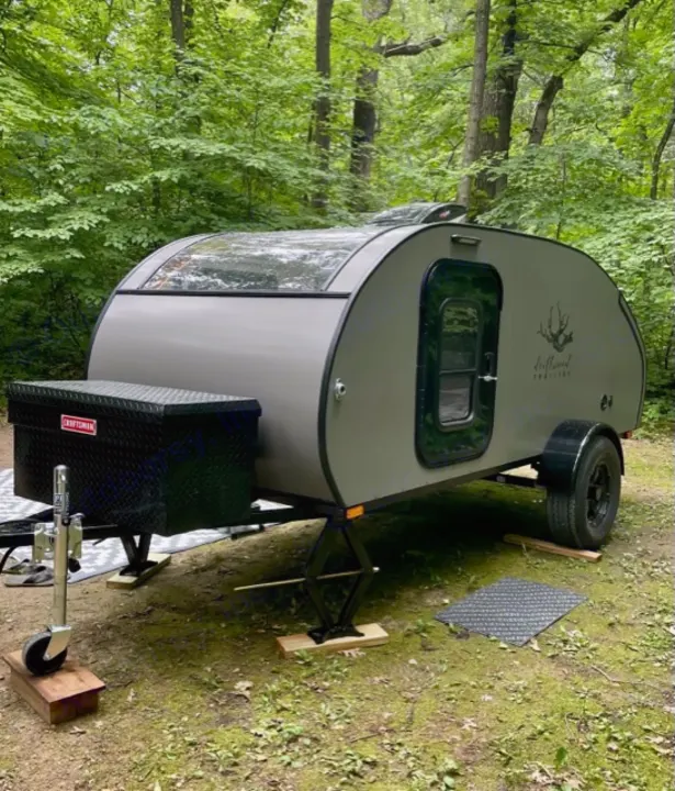 Gray teardrop RV rental, set up with stabilizers, awaits adventure in a lush green forest campsite.. Driftwood Trailers Other 2022