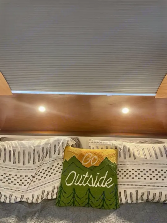 Cozy RV rental bedroom with a 'Go Outside' pillow, wooden headboard, reading lights, and pleated window blinds above.. Driftwood Trailers Other 2022
