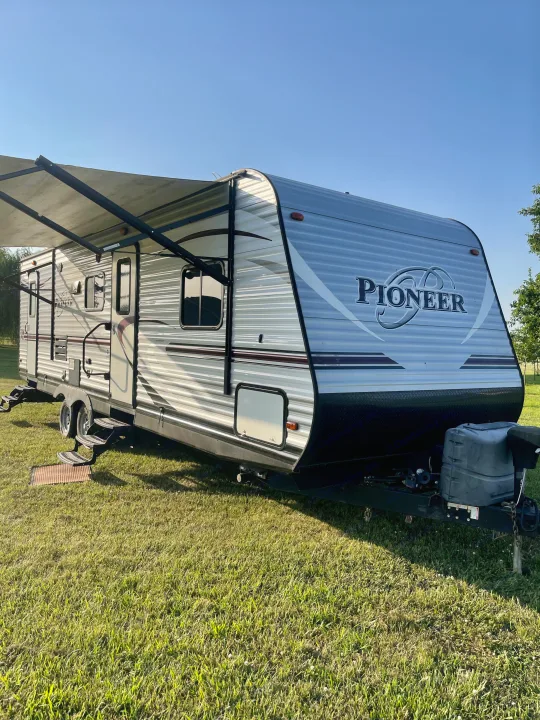 This RV rental, a light-colored Pioneer travel trailer, sits on bright green grass with its awning extended, under a clear blue sky.. Heartland RVs Pioneer 2016