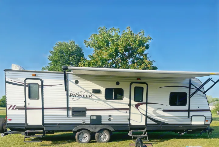 White and grey Pioneer travel trailer RV rental, with an extended awning, parked on grass under a clear blue sky.. Heartland RVs Pioneer 2016