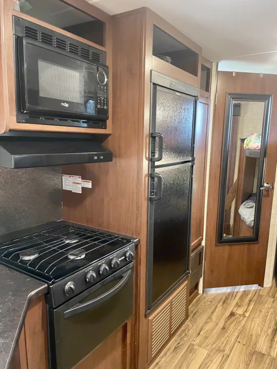 RV rental kitchen featuring a black microwave, gas stove, refrigerator, wood cabinets, and a reflected bunk bed.. Heartland RVs Pioneer 2016