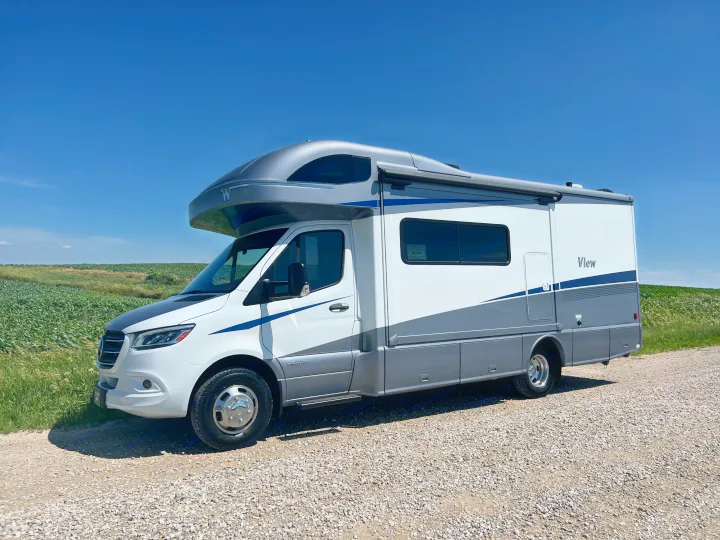 Stylish white & gray Winnebago View RV rental on a gravel road by green fields under blue sky. Perfect for travel.. Winnebago View 2023