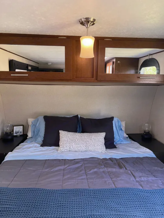 Cozy RV rental bedroom featuring a made bed, side lamps, overhead cabinets, and a central ceiling light.. PTX Forest River PDX170 2019