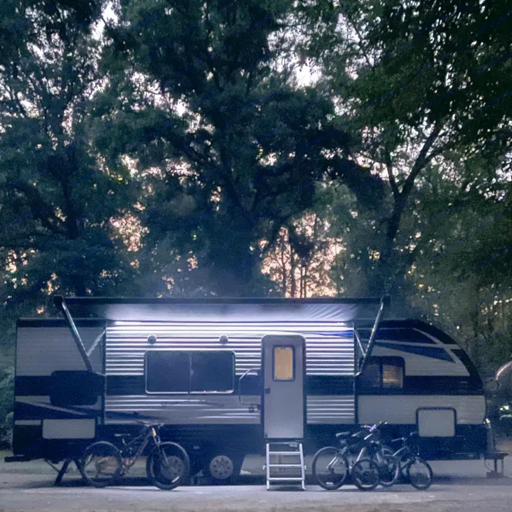 Night camping with an RV rental! Illuminated travel trailer, bikes, and trees under a dark sky. Perfect for outdoor adventure.. Heartland Prowler 2021